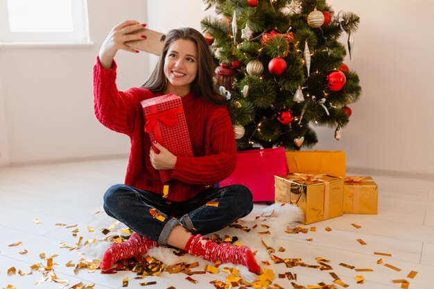 Uśmiechnięta podekscytowana ładna kobieta w czerwonym swetrze siedzi w domu na choince, rozpakowując prezenty i pudełka na prezenty, robiąc zdjęcie selfie aparatem w telefonie