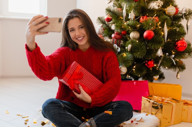 Uśmiechnięta podekscytowana ładna kobieta w czerwonym swetrze siedzi w domu na choince, rozpakowując prezenty i pudełka na prezenty, robiąc zdjęcie selfie aparatem w telefonie