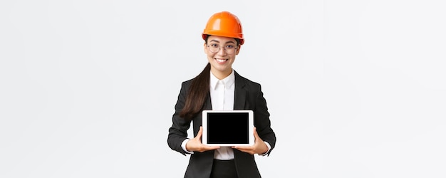 Uśmiechnięta profesjonalna azjatycka architektka przedstawia swojego inżyniera projektu pokazującego schemat na cyfrowym tablecie, który przedstawia prezentację w przedsiębiorstwie lub fabryce stojącej na białym tle