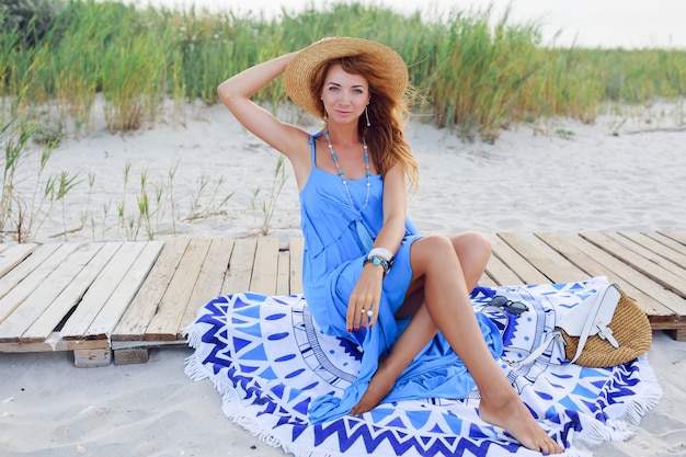 Bezpłatne zdjęcie uśmiechnięta ruda kobieta siedzi na ręczniku plażowym. idealne ciało tan. niebieska sukienka. wietrzne włosy.