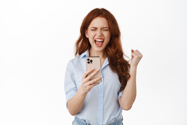 Uśmiechnięta ruda kobieta trzymająca smartfon na białym tle
