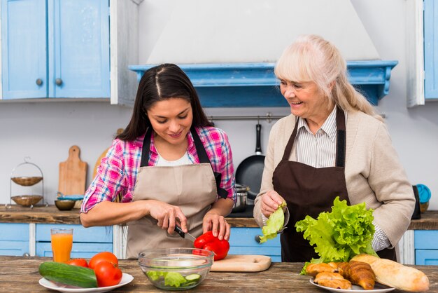 Uśmiechnięta starsza kobieta patrzeje córki tnącego warzywa w kuchni