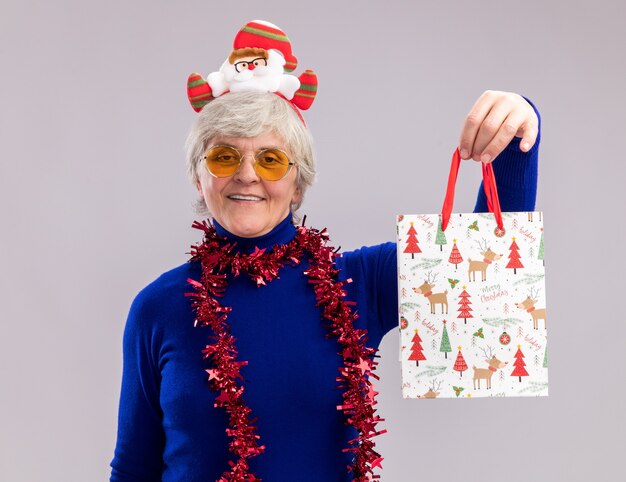 Uśmiechnięta starsza kobieta w okularach przeciwsłonecznych z opaską na głowę Świętego Mikołaja i girlandą wokół szyi trzymającą papierową torbę prezentową odizolowaną na białej ścianie z miejscem na kopię