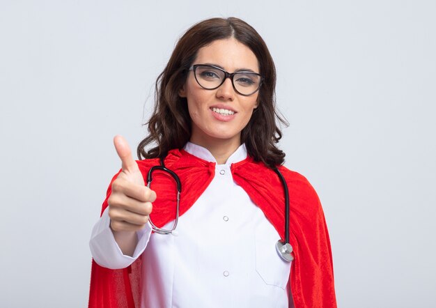 Uśmiechnięta superwoman w mundurze lekarza z czerwoną peleryną i stetoskopem w okularach optycznych kciuki do góry na białym tle na białej ścianie