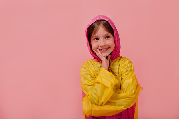 Uśmiechnięta szczęśliwa dziewczyna ubrana w jasny płaszcz przeciwdeszczowy, uśmiechając się i trzymając rękę w pobliżu twarzy