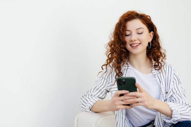 Uśmiechnięta szczęśliwa młoda kobieta z rudymi włosami wysyłająca SMS-y na telefonie leżącym na kanapie w domu