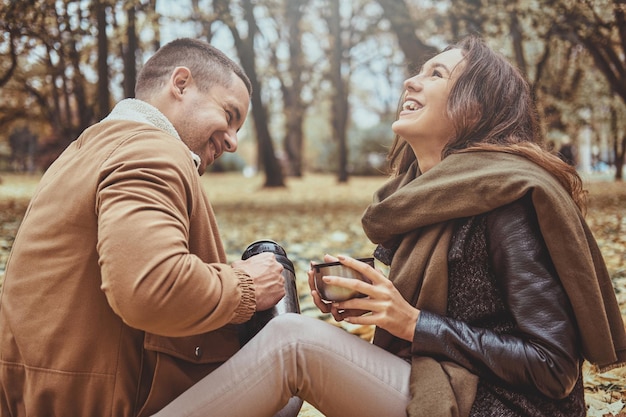 Uśmiechnięta szczęśliwa para pije gorący napój z termosu siedząc w jesiennym parku.