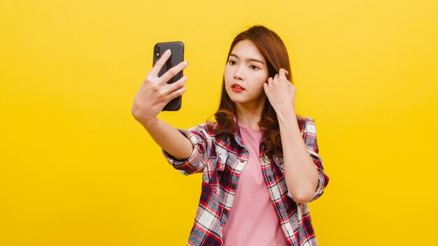 Uśmiechnięta urocza Azjatycka kobieta robi selfie fotografii na smartphone z pozytywnym wyrażeniem w przypadkowej odzieży i patrzeje kamerę nad kolor żółty ścianą. Szczęśliwa urocza uradowana kobieta cieszy się sukcesem.