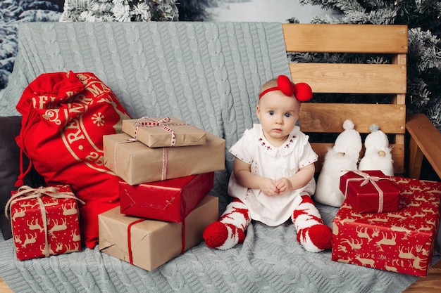 Uśmiechnięta urocza dziewczynka w ślicznej sukni z pałąkiem na głowę siedzi na ławce z dużą ilością świątecznych prezentów