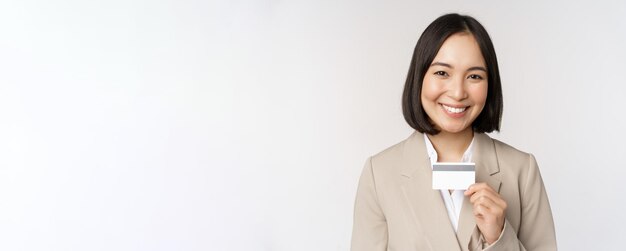 Uśmiechnięta urzędniczka biurowa azjatycka kobieta korporacyjna pokazująca kartę kredytową stojącą na białym tle w beżowym garniturze