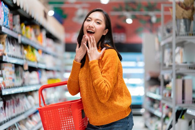 Uśmiechnięta wesoła i radosna kobieta kobieta ręka trzyma koszyk na zakupy gest ręki powitanie niespodzianka stojąca między supermarketem półka z produktem sklep spożywczy supermarket supermarket centrum handlowe