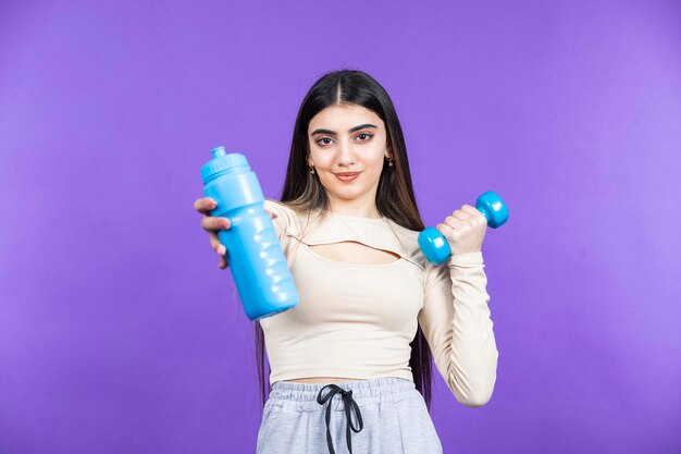 Bezpłatne zdjęcie uśmiechnięta wysportowana dziewczyna pokazująca butelkę wody przed kamerąmłoda wysportowana dziewczyna stoi na fioletowym tle