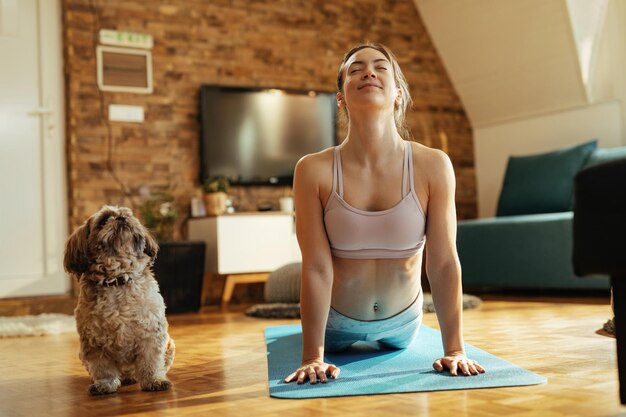Uśmiechnięta wysportowana kobieta w pozie kobry ćwiczy jogę z psem w domu