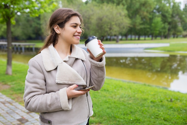 Uśmiechnięta zrelaksowana dziewczyna z telefonem pije smakowitą kawę