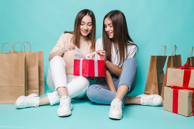 Uśmiechnięte młode dwie dziewczyny siedzą torby na zakupy podłogowe i otwierają prezenty na turkusowej ścianie.