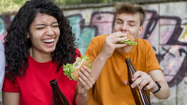 Uśmiechnięte nastolatki jedzą hamburgery na świeżym powietrzu z napojem