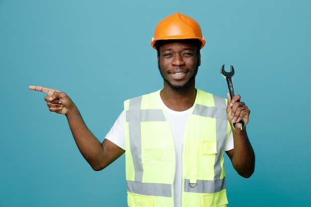 Uśmiechnięte punkty z boku młodego afroamerykańskiego budowniczego w mundurze trzymającego klucz z otwartym końcem na białym tle na niebieskim tle