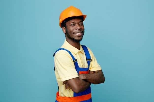 Uśmiechnięte skrzyżowanie rąk młodego afroamerykańskiego budowniczego w mundurze na białym tle na niebieskim tle