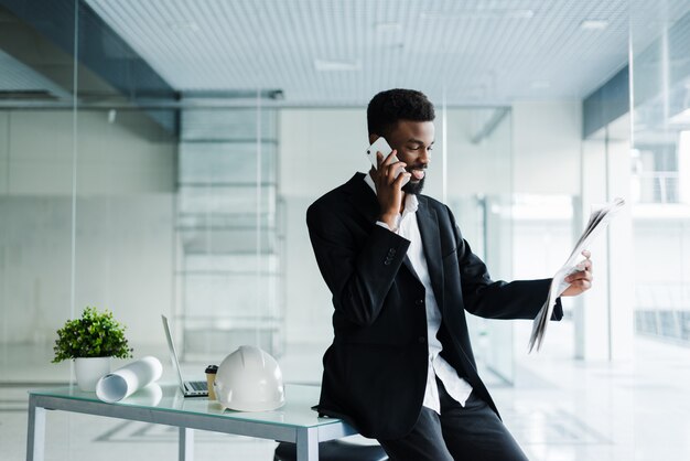 Uśmiechnięty amerykanina afrykańskiego pochodzenia biznesmen opowiada na telefonie z gazetą i filiżanką kawy w biurze