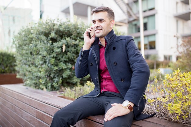 Uśmiechnięty biznesmen opowiada na telefonie komórkowym na ławce