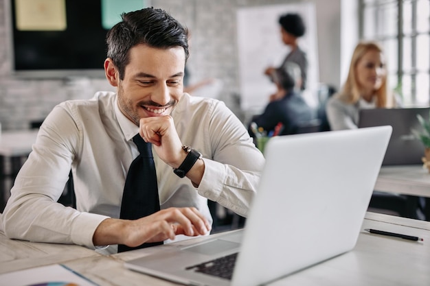 Uśmiechnięty biznesmen siedzi przy biurku w biurze i pracuje na laptopie W tle są ludzie