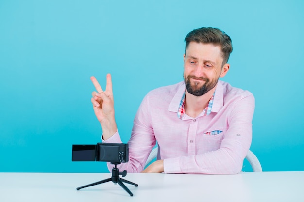 Uśmiechnięty blogger pozuje do swojej mini kamery, pokazując dwa gesty na niebieskim tle