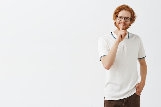 Uśmiechnięty brodaty rudy facet pozuje przy białej ścianie w okularach