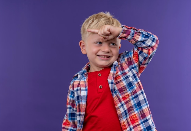Bezpłatne zdjęcie uśmiechnięty chłopiec o blond włosach, ubrany w kraciastą koszulę, wskazujący na coś palcem wskazującym, patrząc z boku na fioletową ścianę