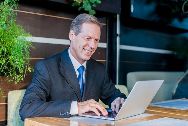 Uśmiechnięty dojrzały biznesmen pracuje na laptopie nad biurkiem w restauracji
