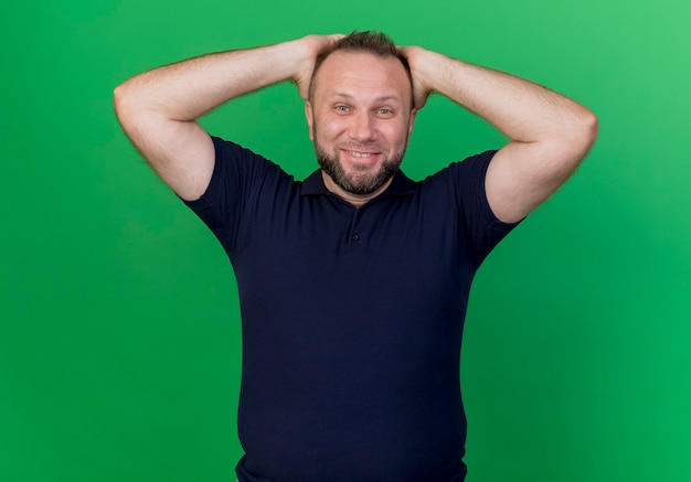 Bezpłatne zdjęcie uśmiechnięty dorosły mężczyzna słowiański wkładając ręce na głowę patrząc na białym tle na zielonej ścianie