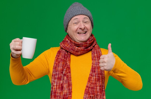 Uśmiechnięty dorosły słowiański mężczyzna w czapce zimowej i szaliku na szyi, trzymający filiżankę i trzymający kciuki w górę