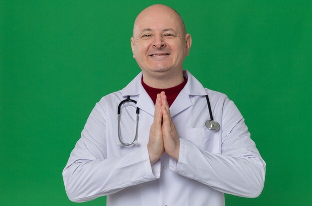 Uśmiechnięty dorosły słowiański mężczyzna w mundurze lekarza ze stetoskopem trzymającym ręce razem