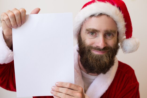 Uśmiechnięty facet jest ubranym Santa kostium i pokazuje pustego papieru prześcieradło