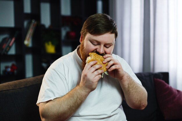 Uśmiechnięty gruby mężczyzna je hamburger siedzi przed telewizorem