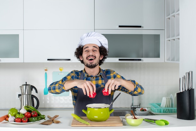 Bezpłatne zdjęcie uśmiechnięty i szczęśliwy męski szef kuchni ze świeżymi warzywami dodający czerwoną paprykę do posiłku w białej kuchni