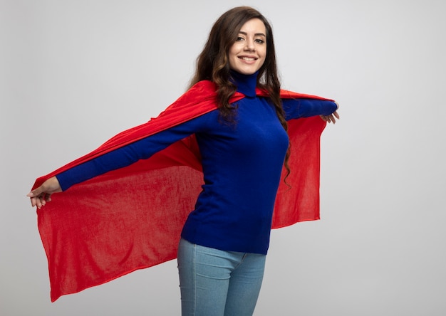 Uśmiechnięty kaukaski dziewczyna superbohatera trzyma czerwoną pelerynę na białym tle