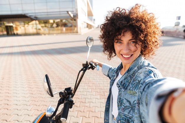 Uśmiechnięty Kędzierzawy Kobiety Obsiadanie Na Nowożytnym Motobike Outdoors I Robić Selfie