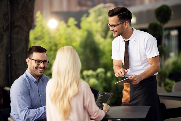 Uśmiechnięty kelner używający touchpada podczas rozmowy z gośćmi i przyjmowania zamówienia