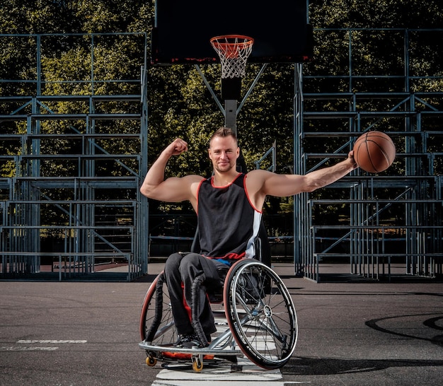 Uśmiechnięty koszykarz kaleka na wózku inwalidzkim trzyma piłkę na otwartym boisku.