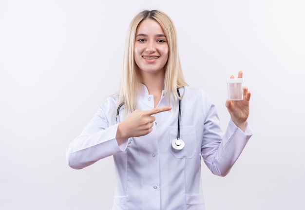 Uśmiechnięty lekarz młoda blondynka ubrana w stetoskop i fartuch medyczny w ortezie dentystycznej wskazuje opróżnić puszkę trzymając ją za rękę na odosobnionym białym tle