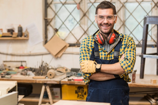 Uśmiechnięty męski cieśla jest ubranym zbawczych szkła stoi przed workbench z jego ręką krzyżującą