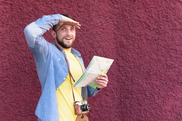 Bezpłatne zdjęcie uśmiechnięty męski podróżnika fotograf osłania jego oczy z mienie mapy pozycją przeciw textured ścianie