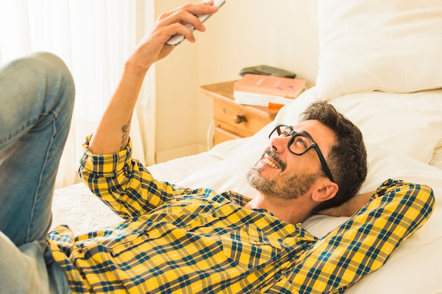 Uśmiechnięty mężczyzna leży na łóżku patrząc na telefon komórkowy