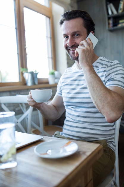 Uśmiechnięty mężczyzna trzyma kubek kawy i rozmawia przez telefon komórkowy