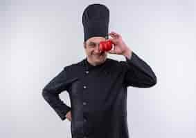 Bezpłatne zdjęcie uśmiechnięty mężczyzna w średnim wieku kucharz w mundurze szefa kuchni objęte oko z pieprzem, kładąc rękę na biodrze