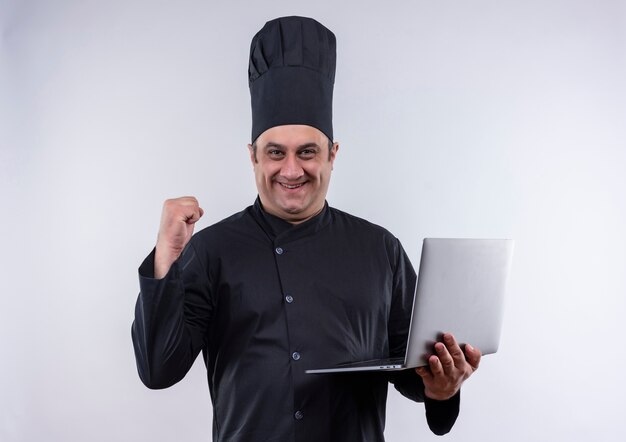 Uśmiechnięty mężczyzna w średnim wieku kucharz w mundurze szefa kuchni trzymając laptob pokazuje tak gest