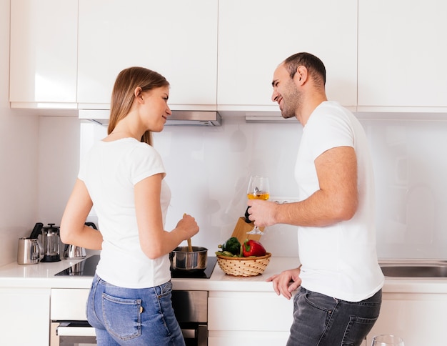 Uśmiechnięty młodego człowieka mienia wineglass w ręce patrzeje jej żony przygotowywa jedzenie w kuchni