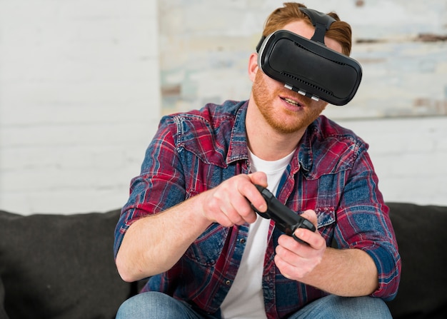 Uśmiechnięty młodego człowieka obsiadanie na kanapie jest ubranym rzeczywistość wirtualna szkła bawić się z joystickiem