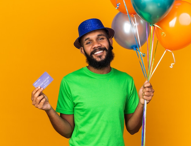 Uśmiechnięty młody afro-amerykański facet w imprezowym kapeluszu, trzymający balony i kartę kredytową
