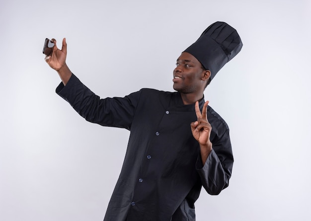 Uśmiechnięty młody afro-amerykański kucharz w mundurze szefa kuchni patrzy na telefon i gesty znak zwycięstwa na białym z miejsca na kopię